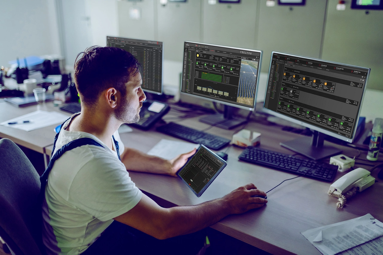 Hardworking plant worker sitting in control room, holding tablet and using computer for monitoring.