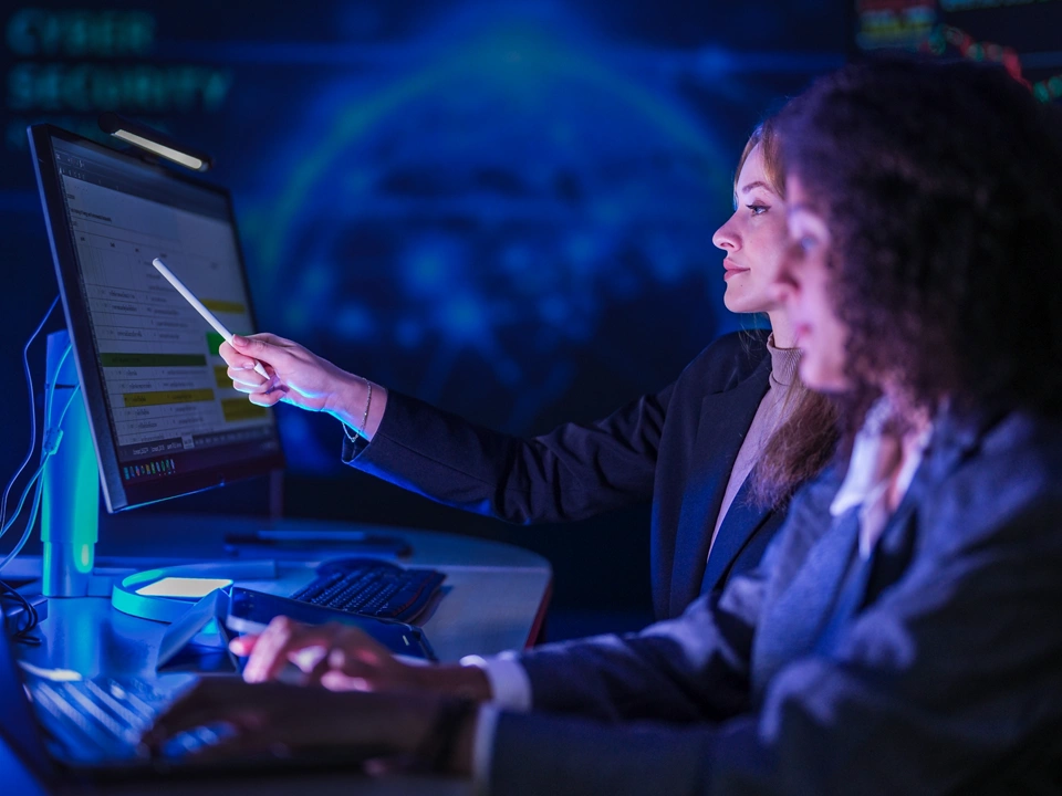 Cyber security specialists monitoring global network threats. Female tech expert pointing at digital system data on screen while partner analyzing code at night.