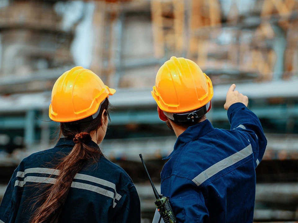Two workers wearing hard hats examine machinery at an industrial facility. One points toward equipment while discussing safety protocols, emphasizing teamwork and attention to detail.