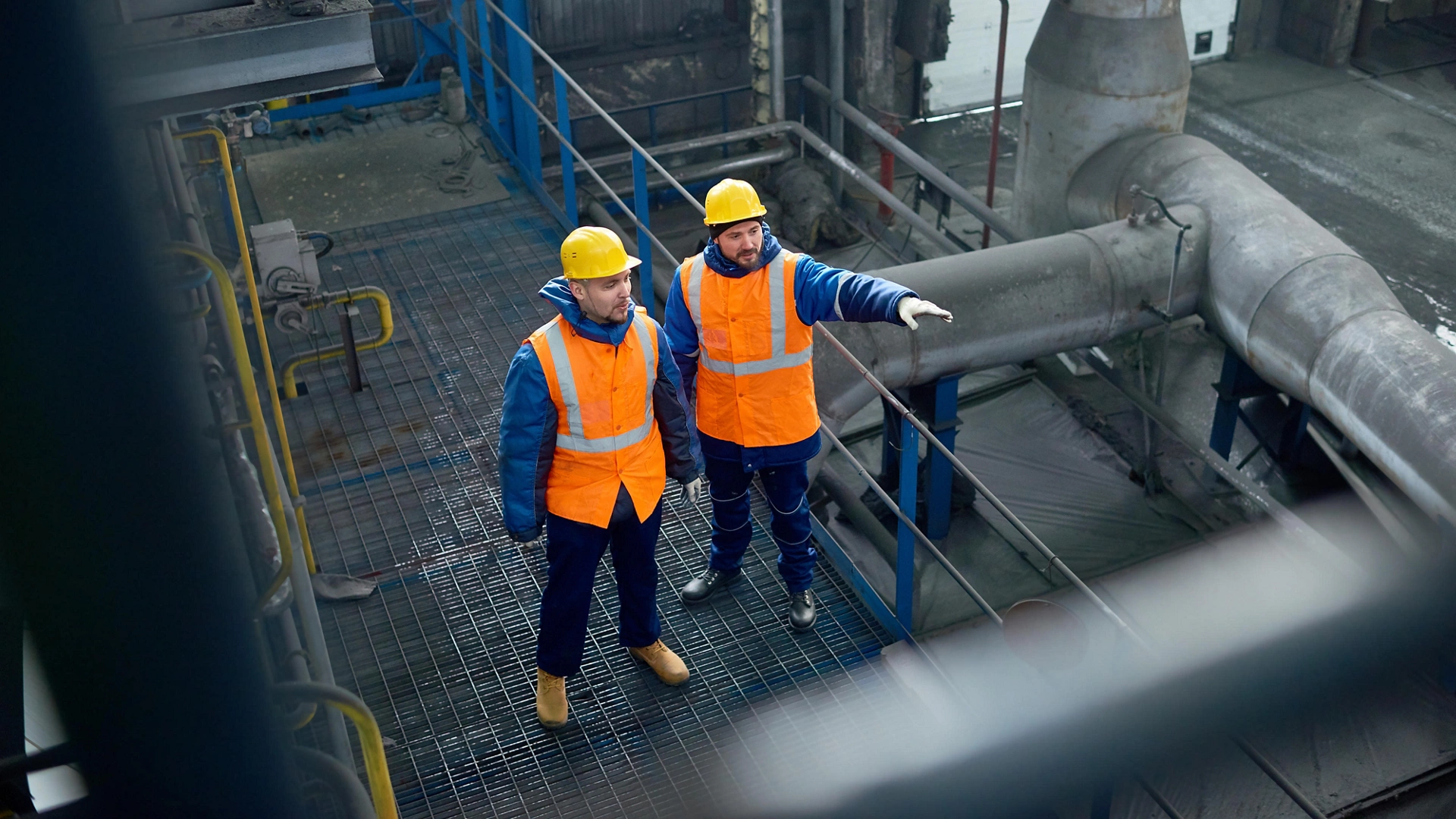 High angle view of bearded engineers wearing uniform and protective helmets standing at spacious production department of modern plant and brainstorming on joint work, High angle view of bearded engineers wearing uniform and protective helmets standing at spacious production department of modern plant and brainstorming on joint work
