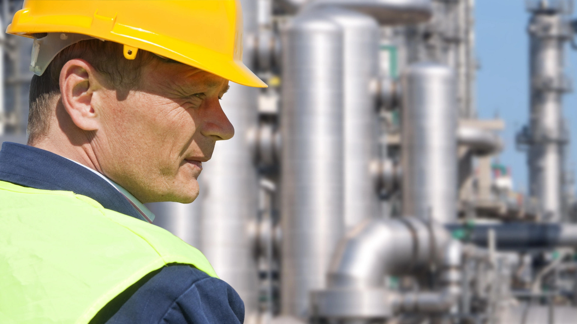Close up of an engineer wearing a safety vest, blue coveralls, and a hard hat in front of a petrochemical plant