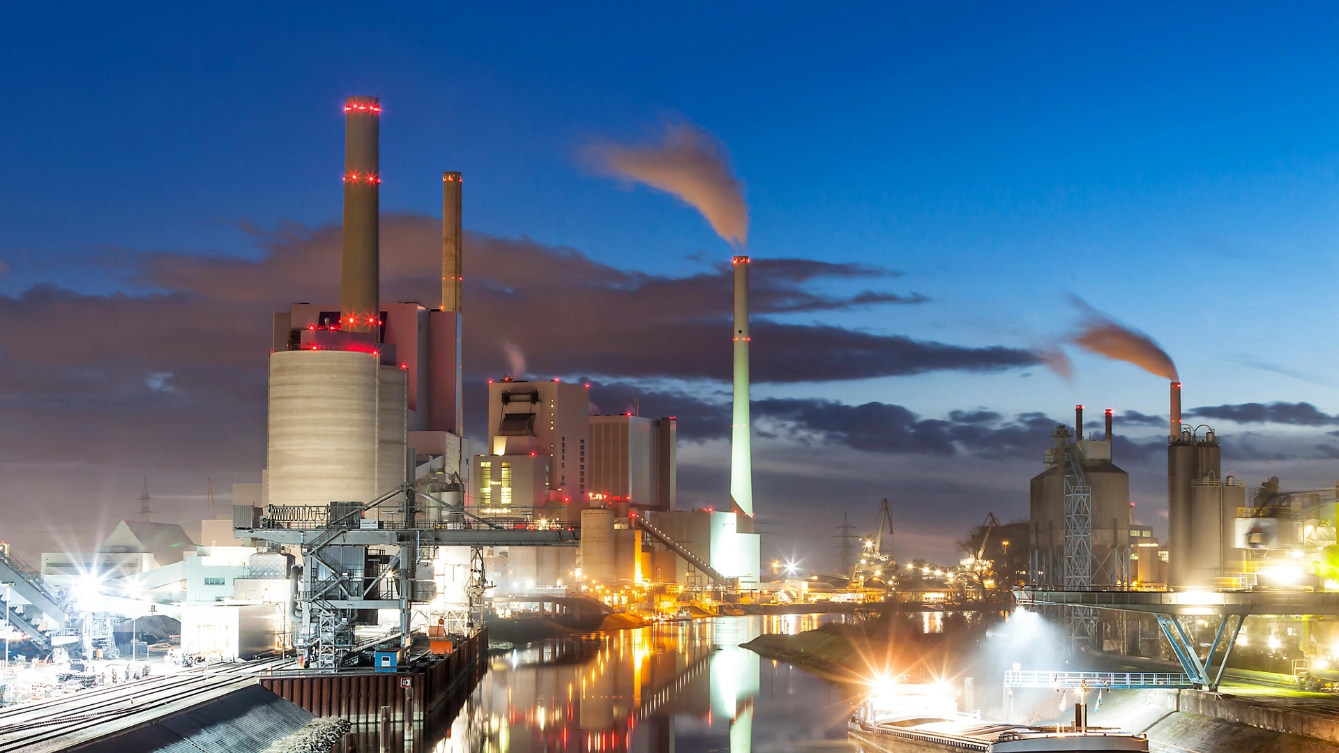 A coal power plant sits at the river's edge at dusk.  Lights from the buildings and streets glow, and a reflection of the whole scene can be seen on the water of the calm river below.