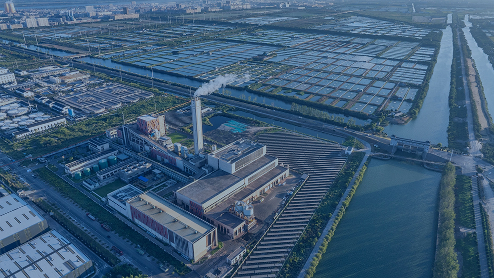 aerial-view-of-industrial-plant-with-smokestack-emitting-steam-surrounded-by-rectangular-water-treatment-basins-canals-and-urban-area-in-background-under-clear-sky