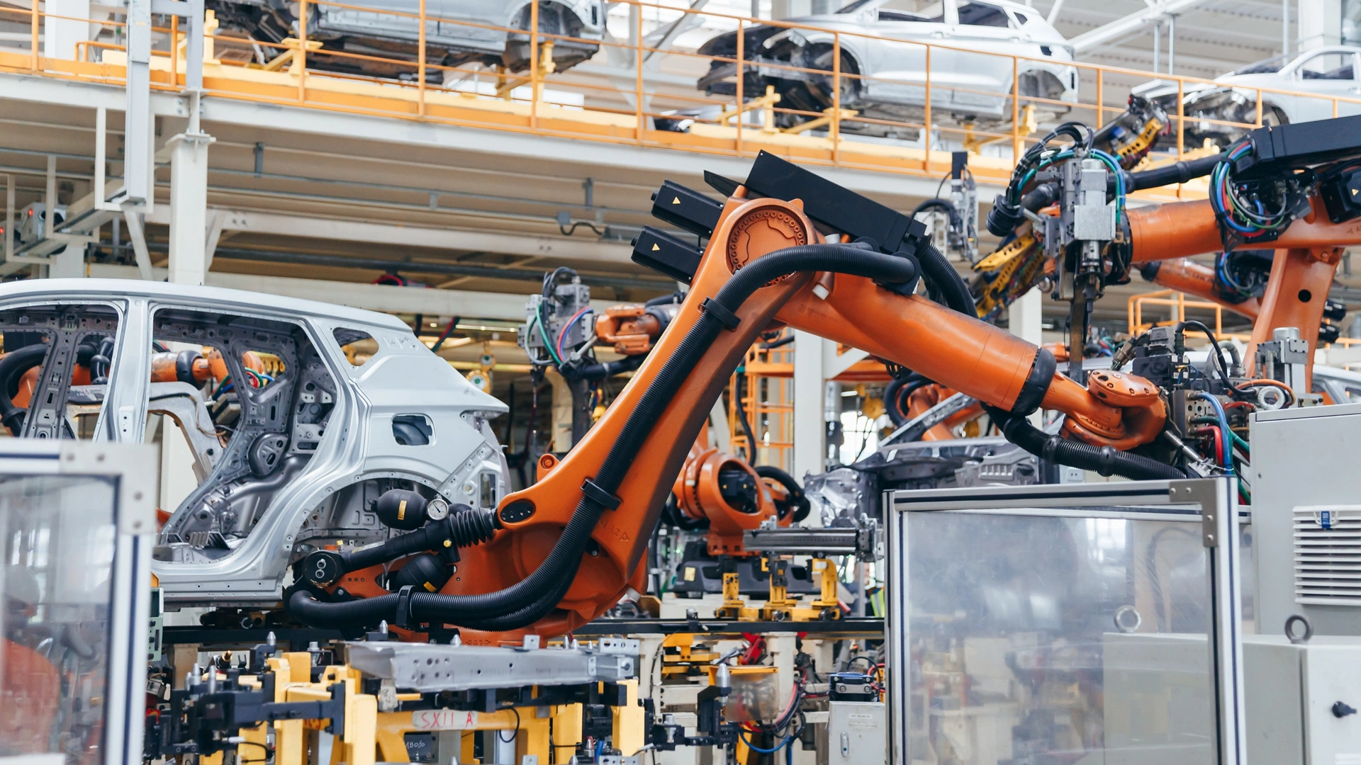 Multiple orange robotic arms welding and working on steel car bodies suspended on elevated platforms