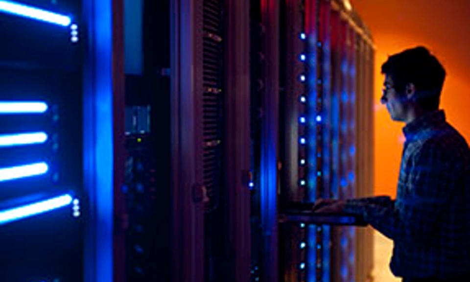 The interior of a modern server room in a data center.  An IT engineer is busy configuring the servers. The room is dark, but the servers themselves are lit.  The servers at the left side are lit in blue, while the ones at the other end are lit by an orange light source coming from the right side.