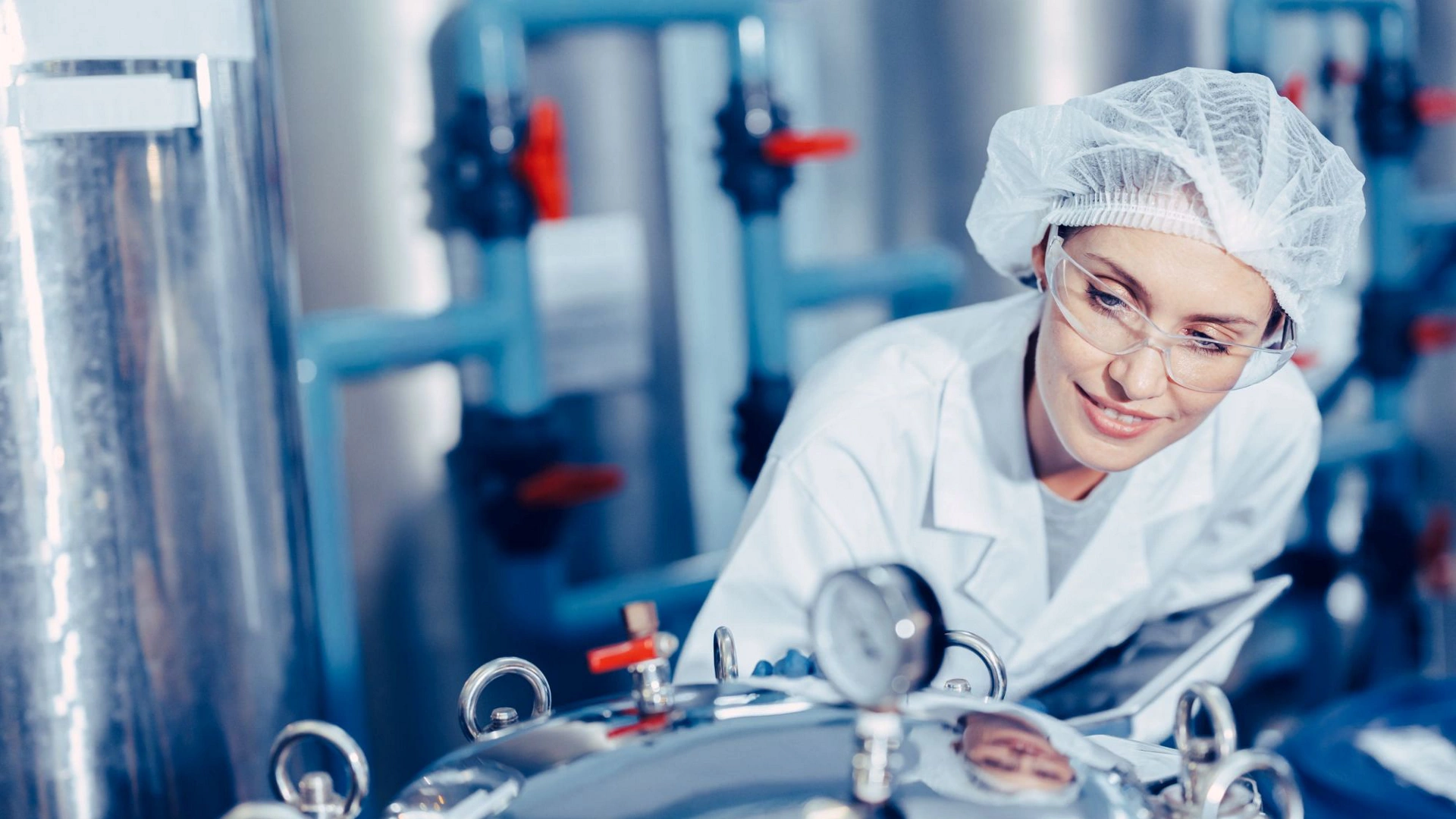 person-wearing-lab-coat-hair-cover-and-safety-glasses-inspecting-stainless-steel-industrial-processing-equipment-with-pressure-gauge-and-piping-in-manufacturing-facility