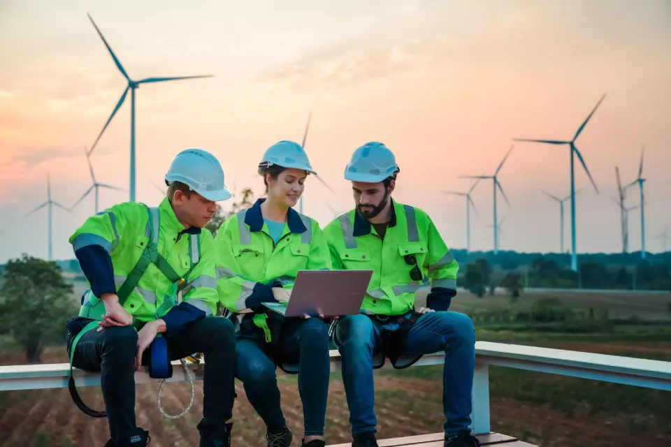 three-wind-energy-workers-in-high-visibility-jackets-and-safety-helmets-reviewing-laptop-data-at-wind-farm-with-multiple-turbines-at-sunset